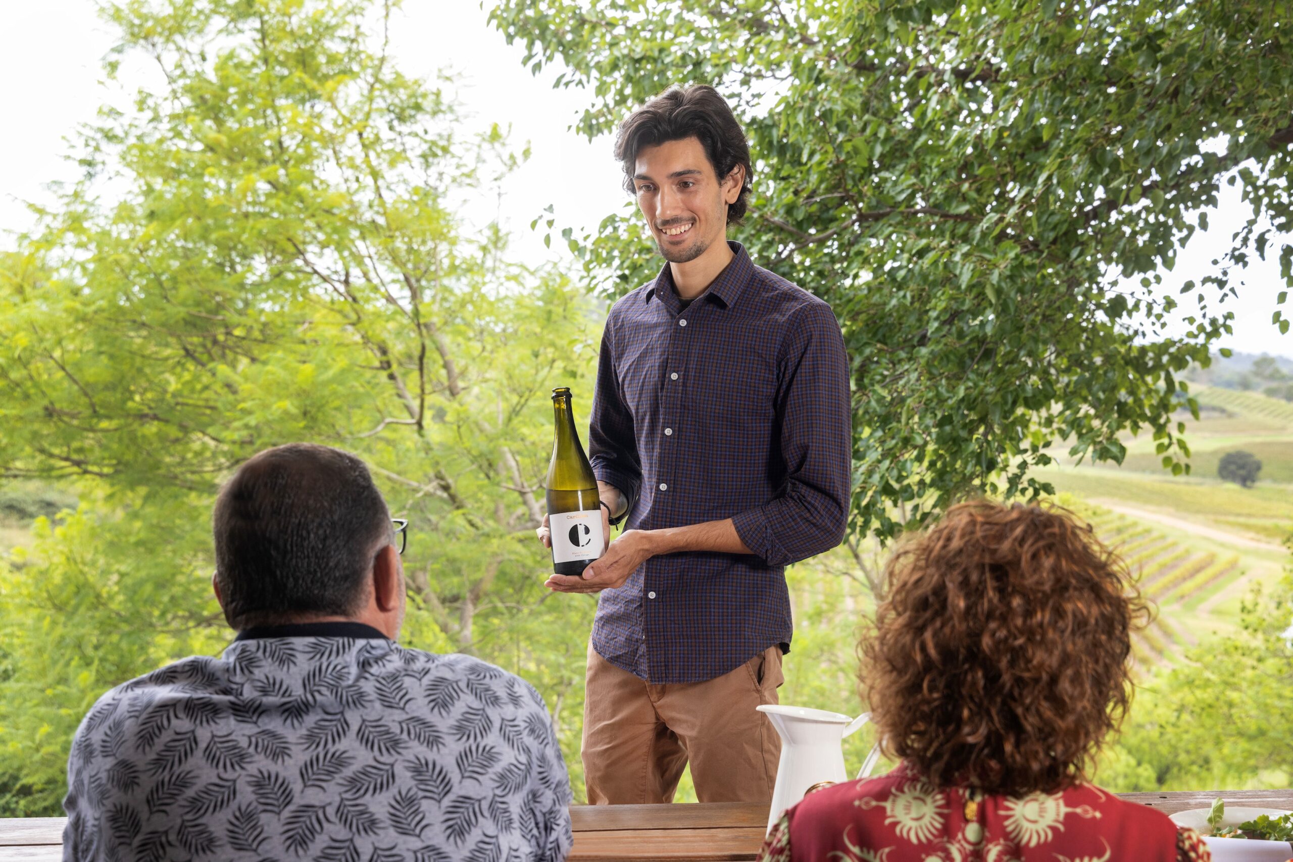 Man serving wine to guests outdoors with scenic valley view in background.